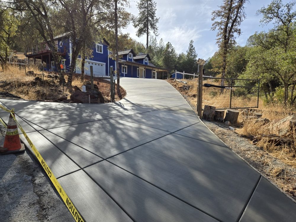 Freshly poured concrete driveway on sloped terrain leading to blue residential home with trees
