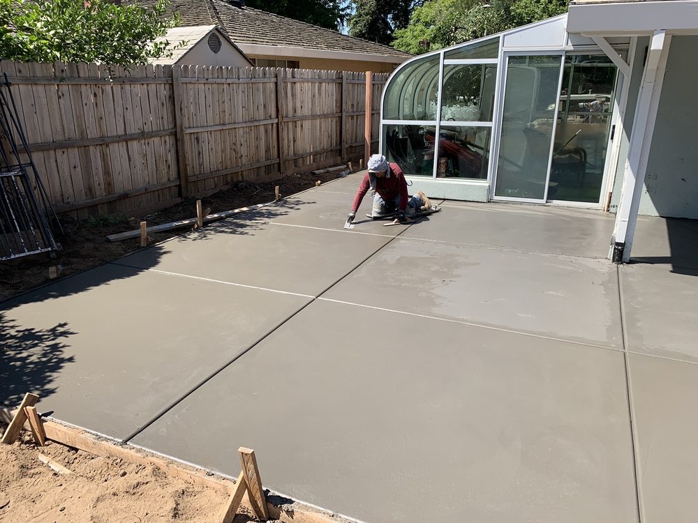 Worker finishing freshly poured concrete patio in residential backyard with wooden fence and sunroom